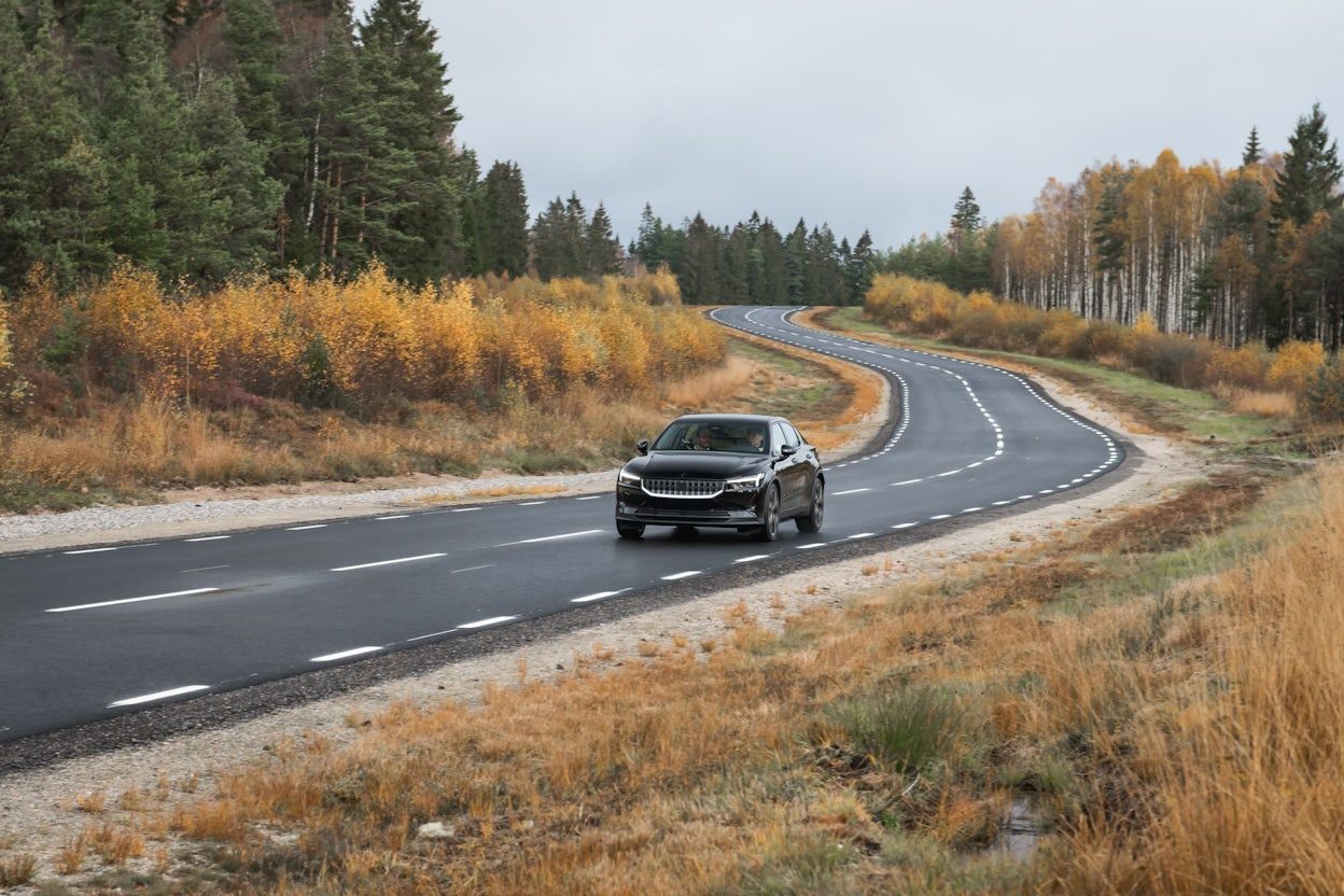 A black Polestar 2 driving on a forest road in autumn.