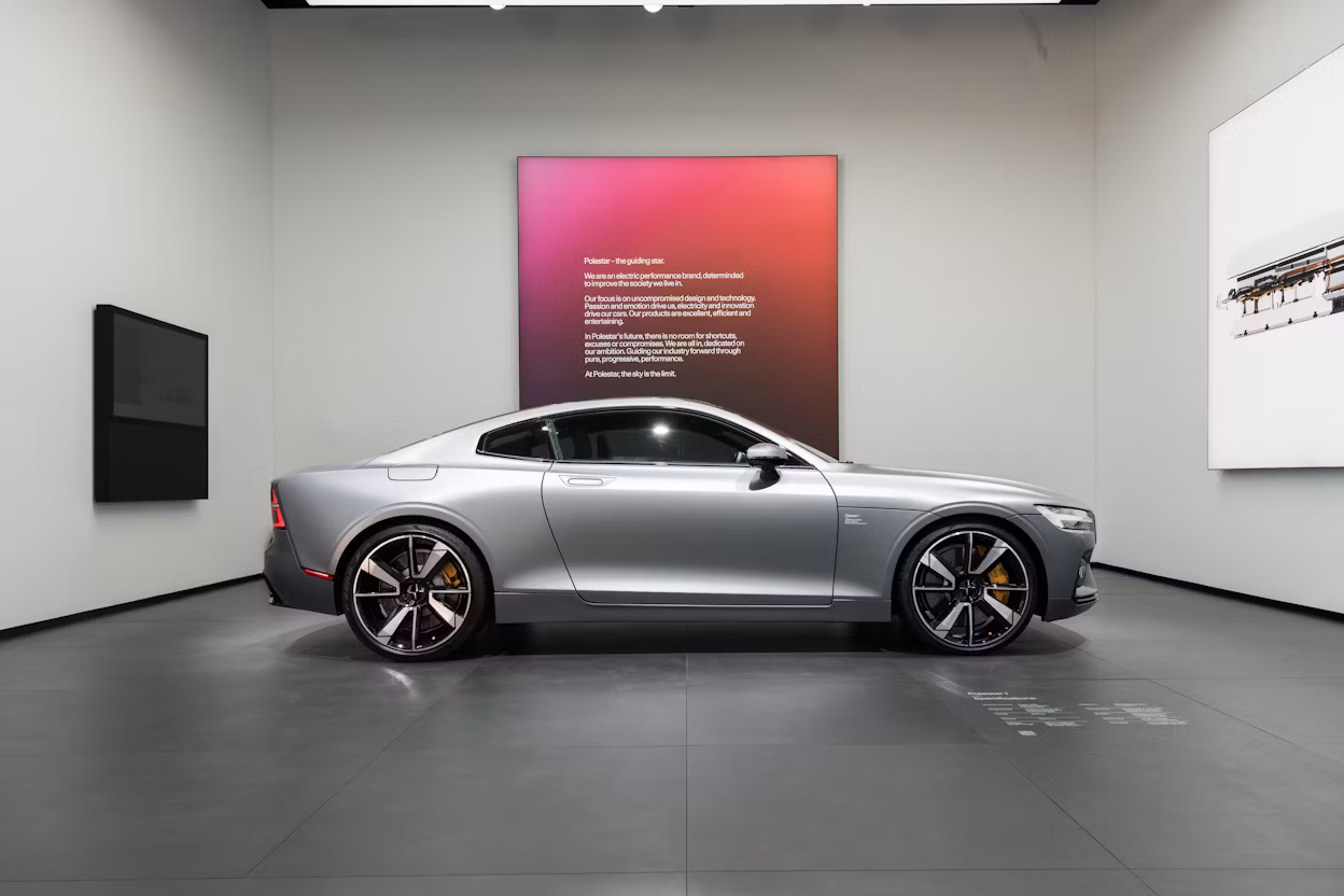 Side view of a silver Polestar 1, positioned in a well-lit exhibition space, with the brand's guidelines prominently showcased on a large screen.