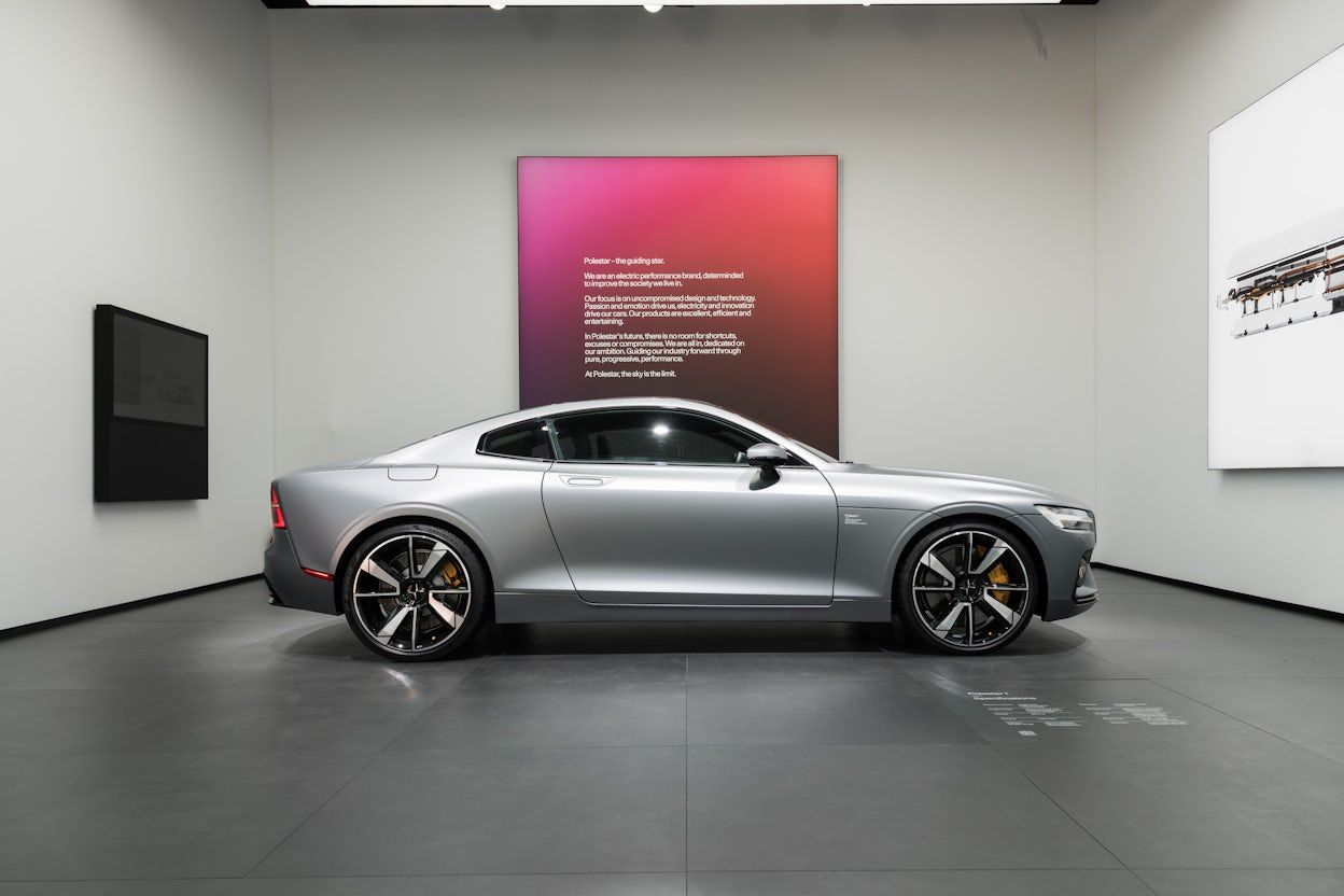 Side view of a silver Polestar 1, positioned in a well-lit exhibition space, with the brand's guidelines prominently showcased on a large screen.