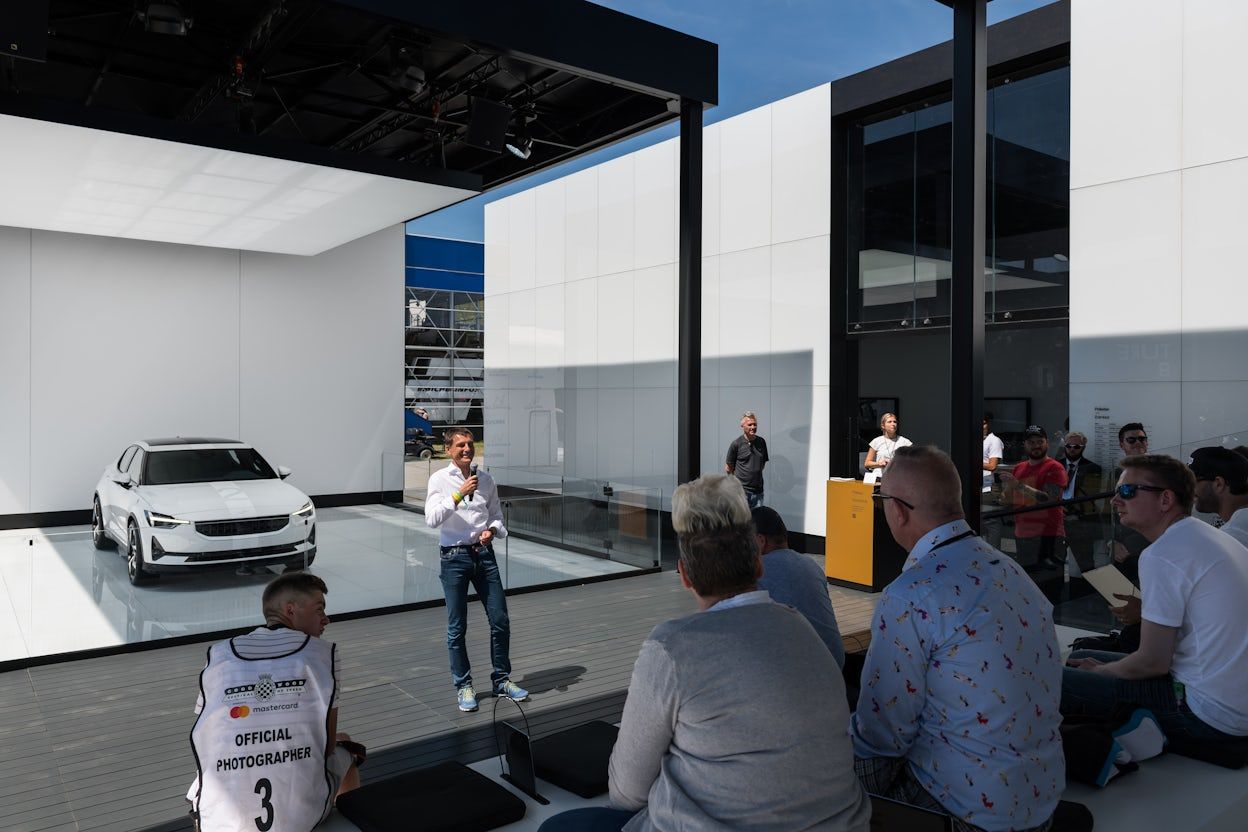 Polestar CEO Thomas Ingenlath speaking in front of a seated crowd, with a white Polestar 2 displayed in the background.