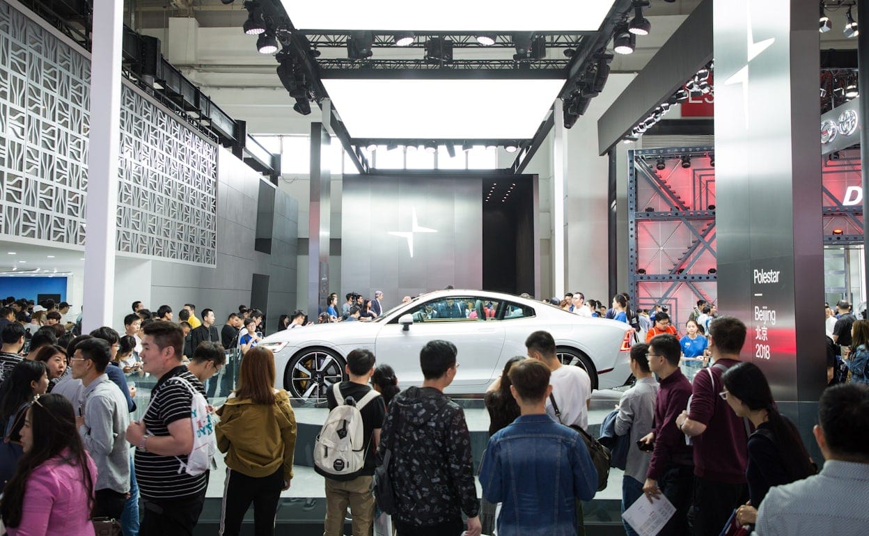 A crowd watching a white Polestar 1 on display at car event, with the Polestar logo evident in the background.