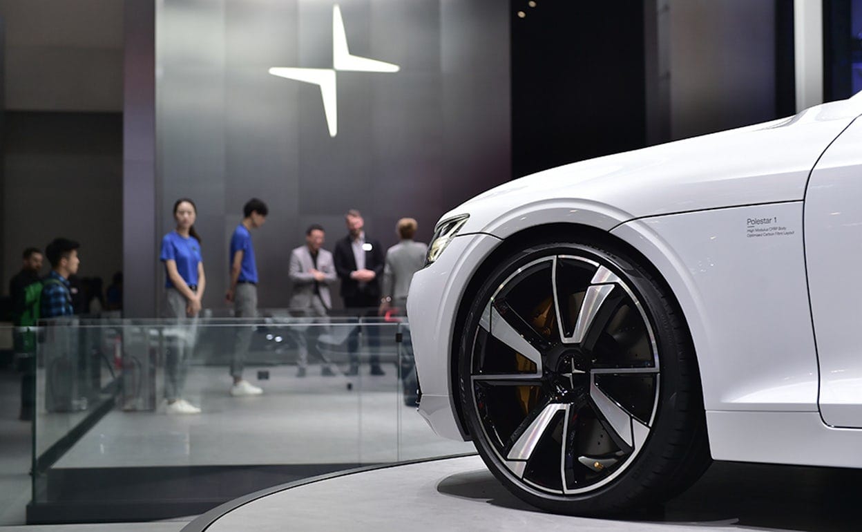 Focus on the left front of a white Polestar 1, with people and the Polestar logo in the background.