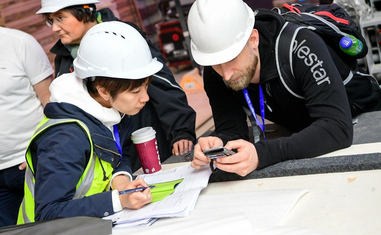 Two people in hard hats leaning against a table and looking at a phone