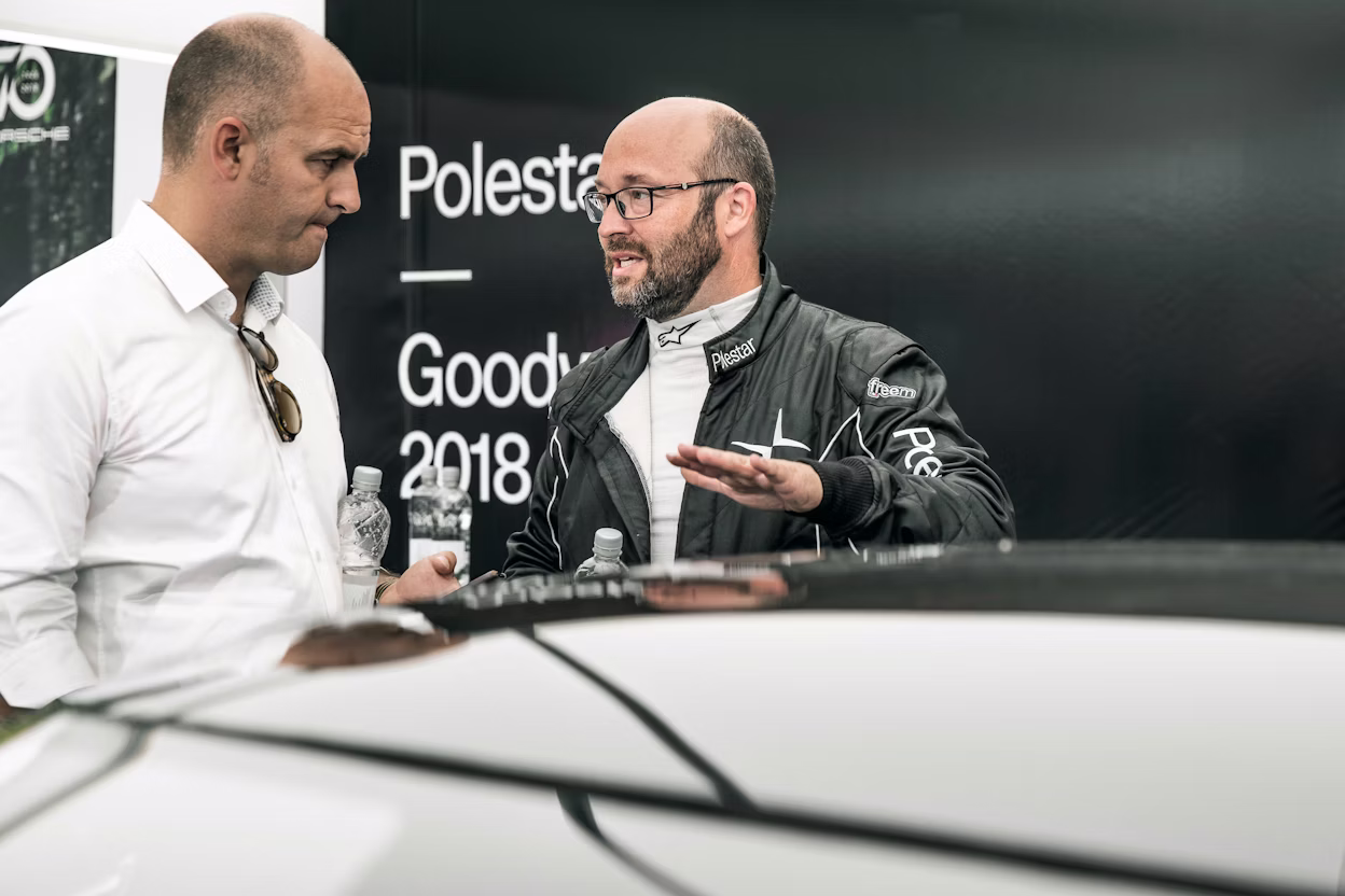A man wearing a Polestar racing suit speaking to another man in a white shirt.
