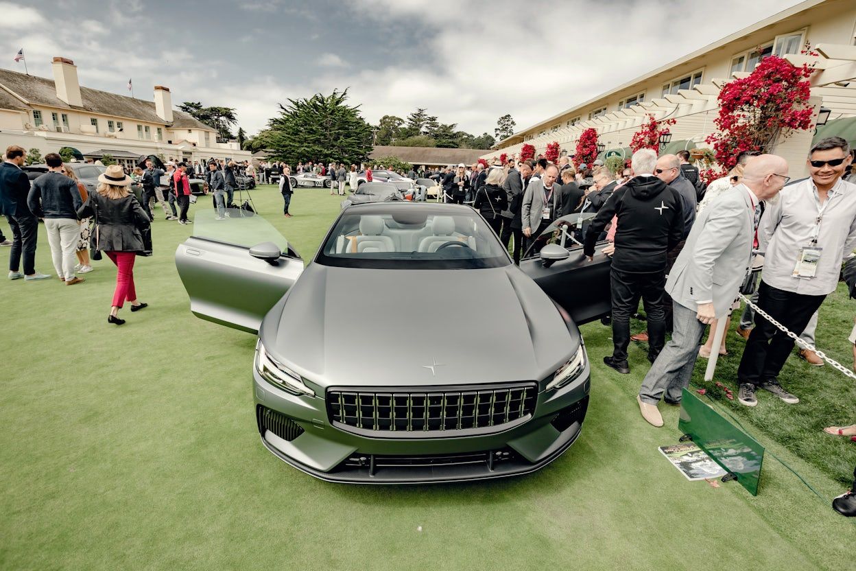 Front view of a metallic Polestar 1 outside a fancy event ground with lots of people and red flowers.