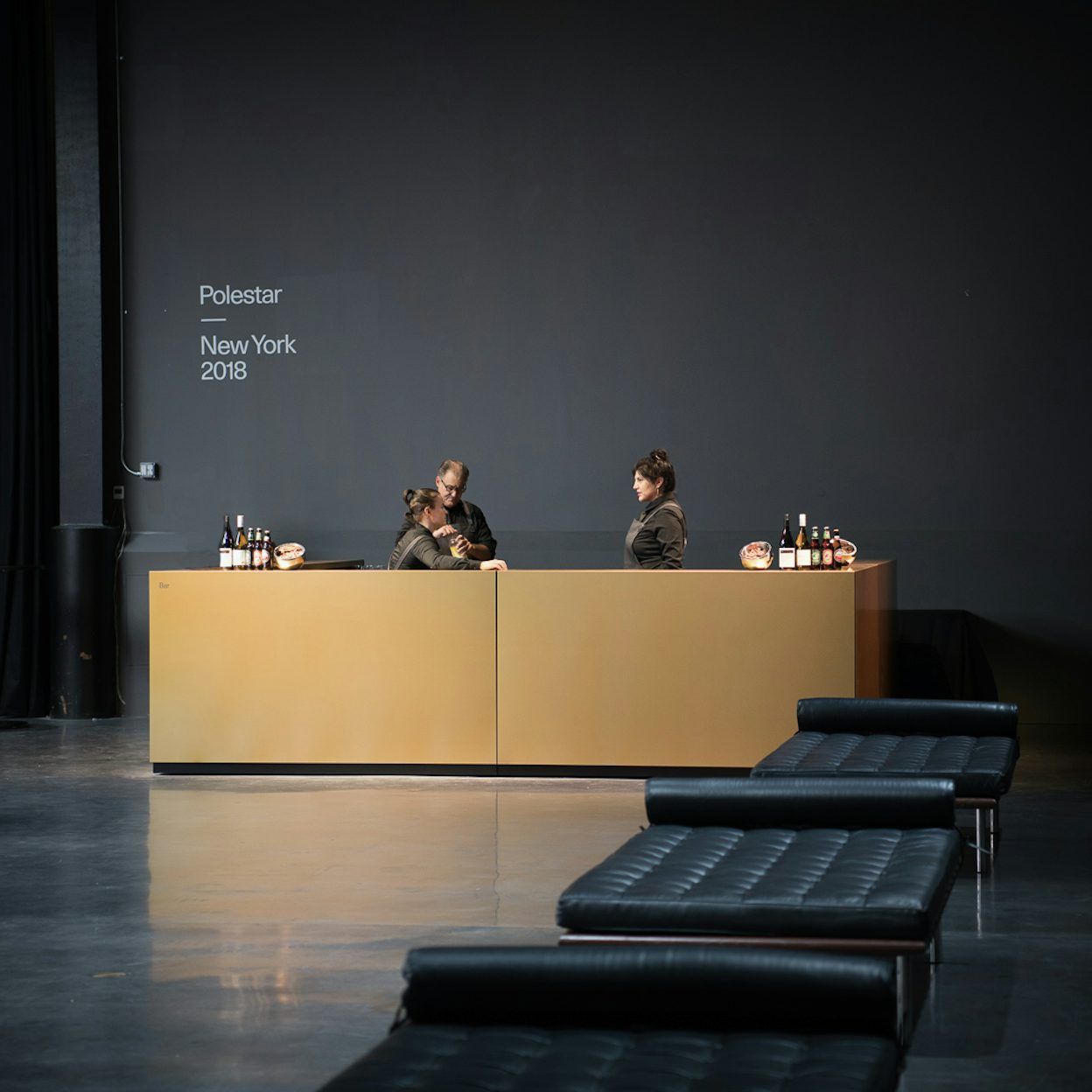 Three people behind a bar in front of a black wall with the text Polestar New York 2018.