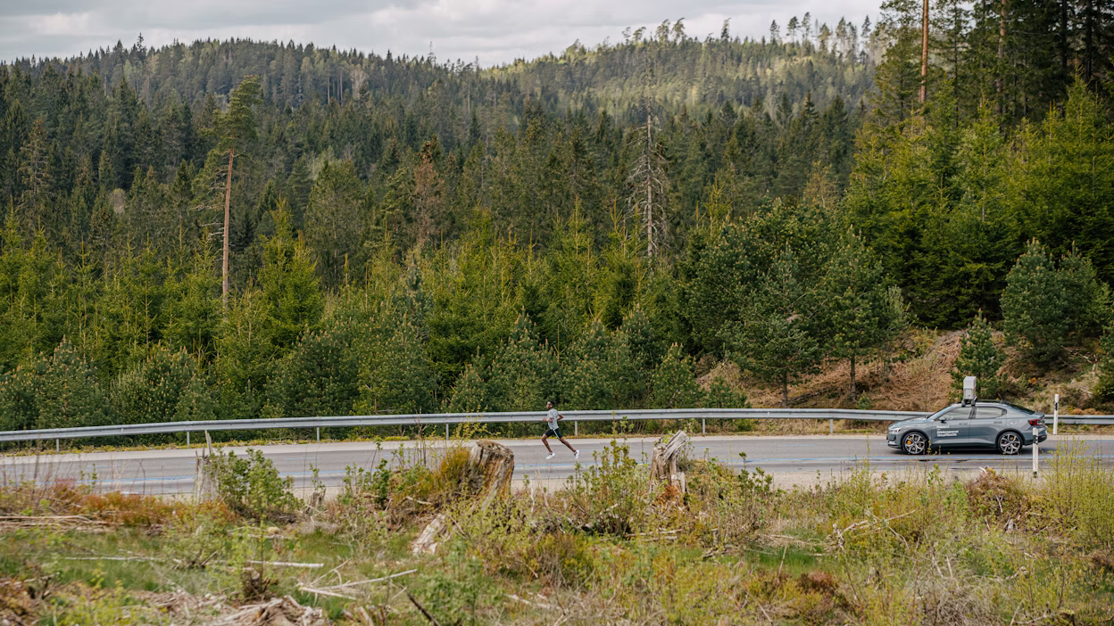 Distant view of a grey Polestar 2 driving on a forest road with a man running in front of it.