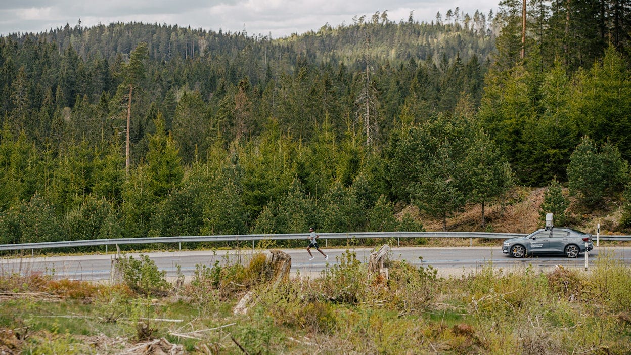 Distant view of a grey Polestar 2 driving on a forest road with a man running in front of it.