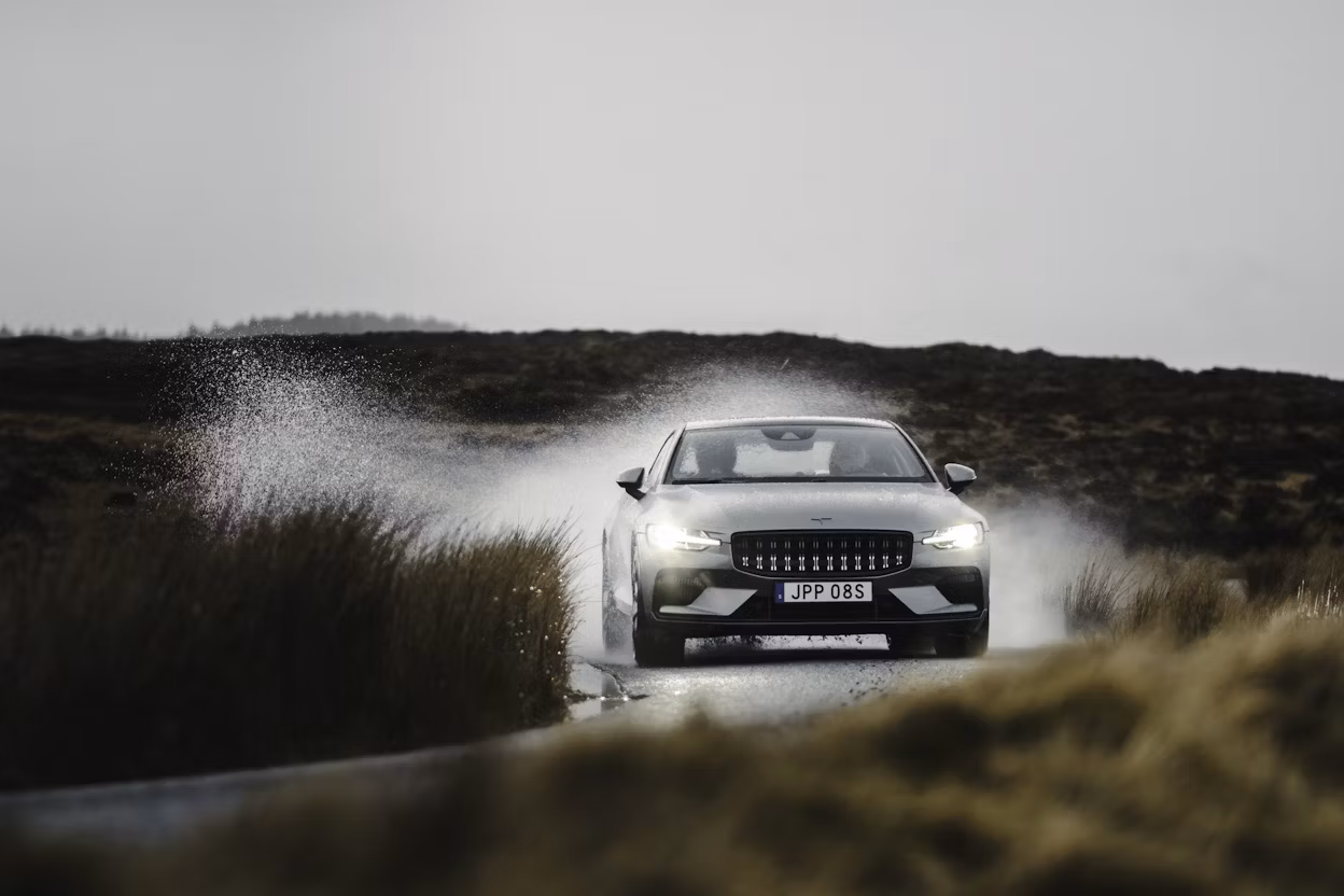 A grey Polestar 1 driving on a wet road surrounded by brown grass.