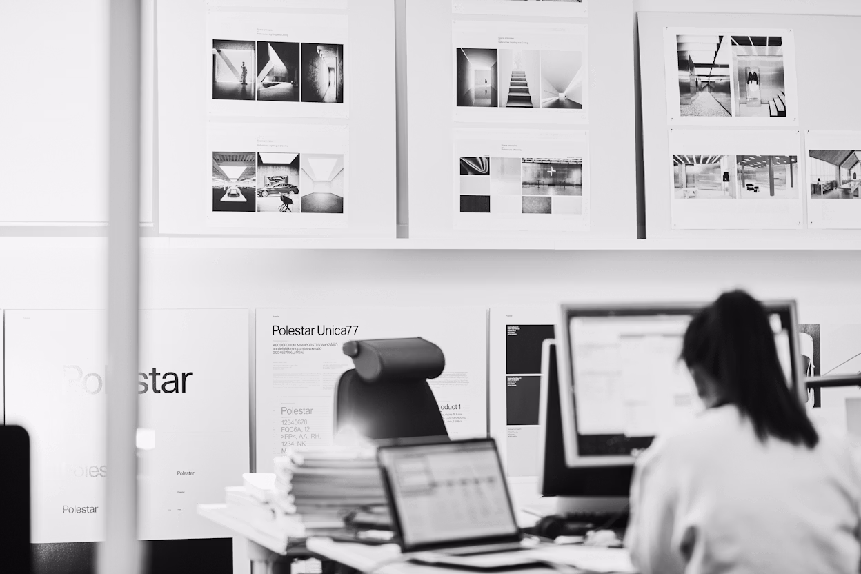 Person working at a desk with computer screens, papers, and Polestar branding on posters and documents in the background.