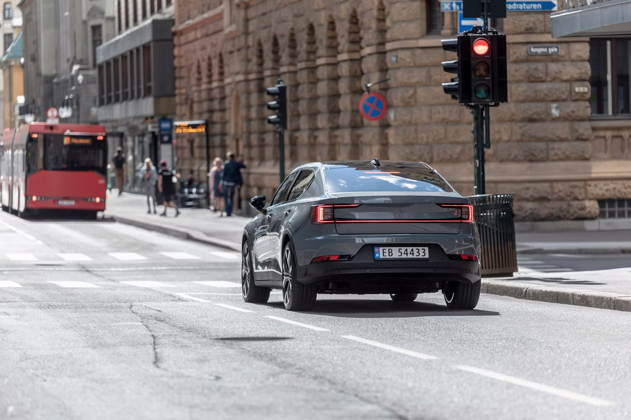 Polestar car driving on an urban street near a red bus, pedestrians, and traffic lights.