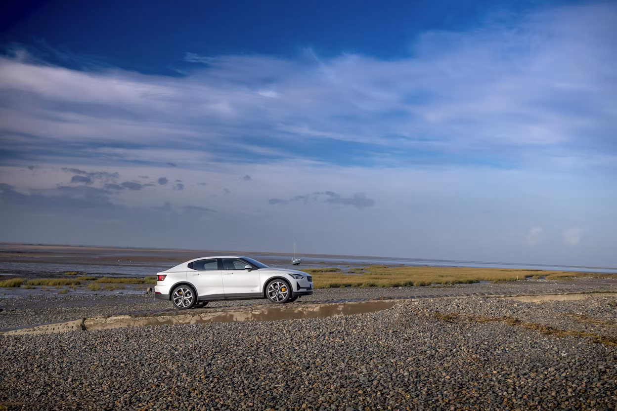 A white Polestar 2 on a stone beach under a blue sky.