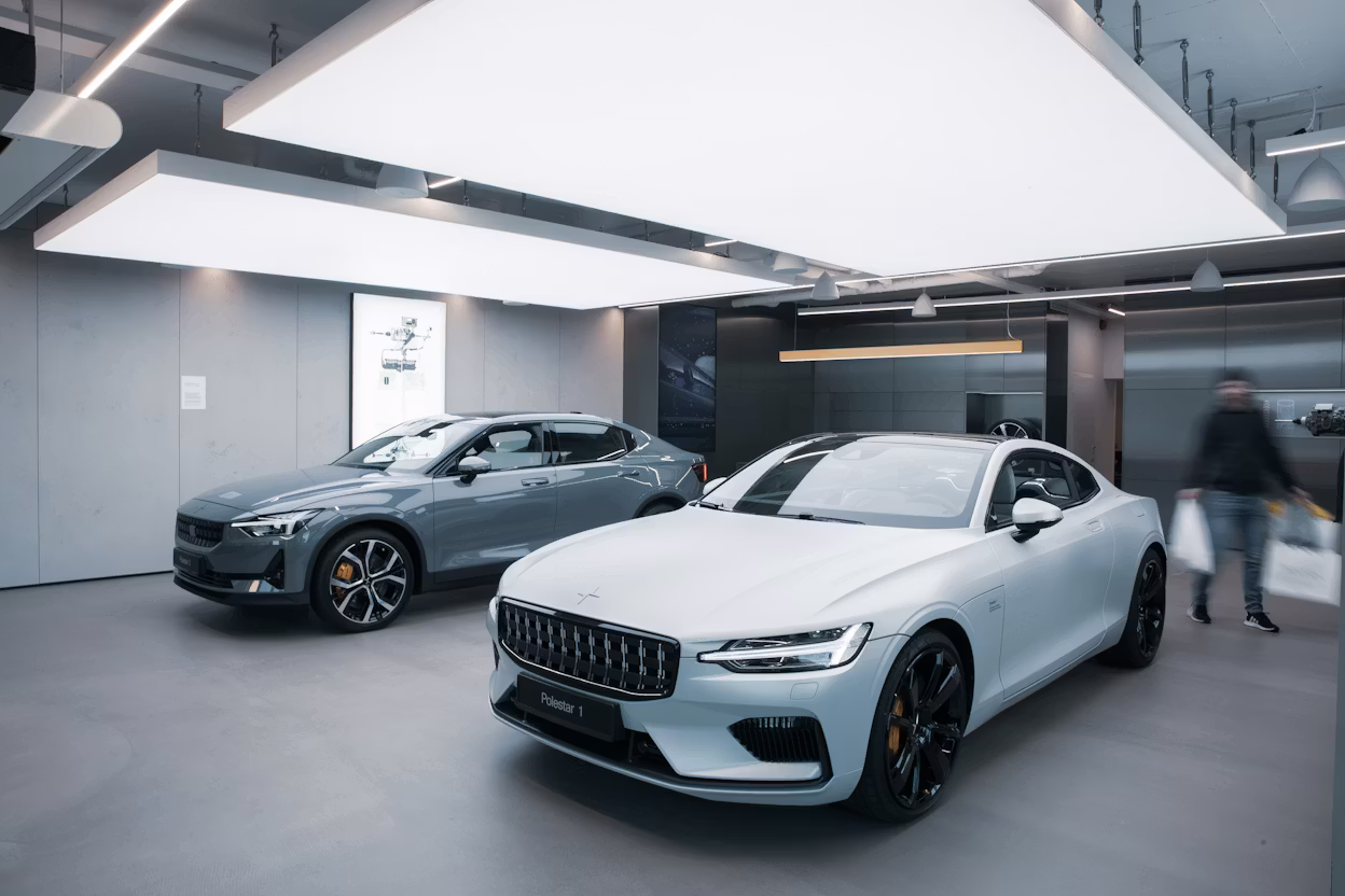 Polestar cars displayed in a showroom with gray walls, bright ceiling lights, and a blurred person walking while holding bags.