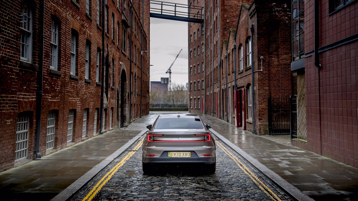 Rear view of a silver Polestar 2 on the road between two brick buildings.