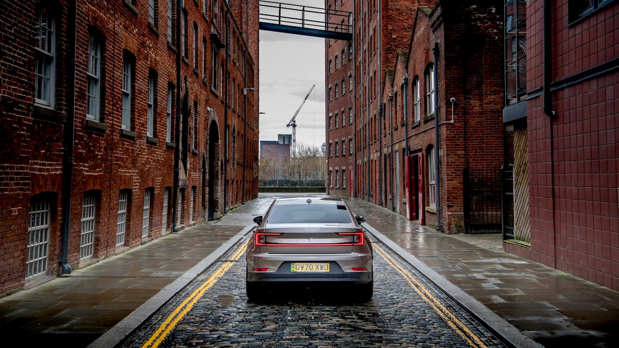 Rear view of a silver Polestar 2 on the road between two brick buildings.