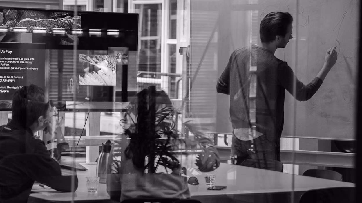 Image captured through a glass wall showing a man writing on a whiteboard and two people watching.