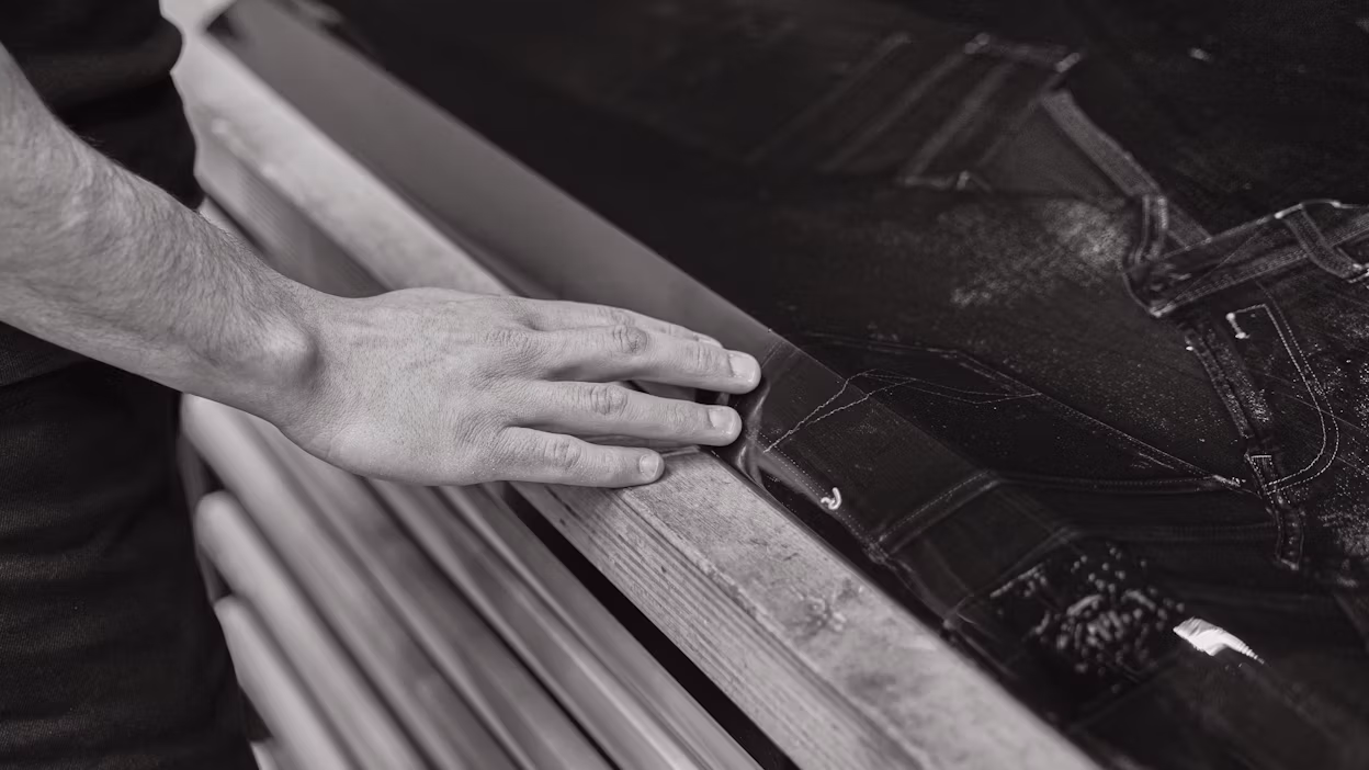 A hand resting on the edge of a wooden surface near a piece of dark fabric with stitching.