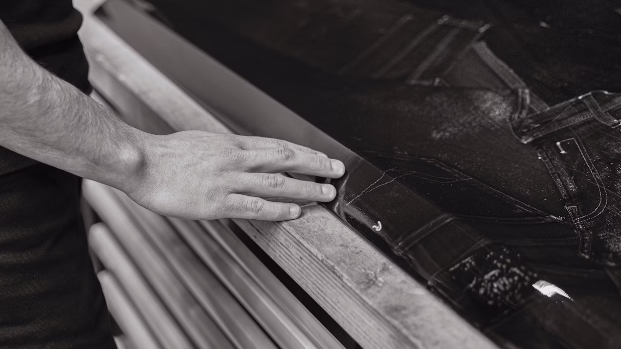 A hand resting on the edge of a wooden surface near a piece of dark fabric with stitching.