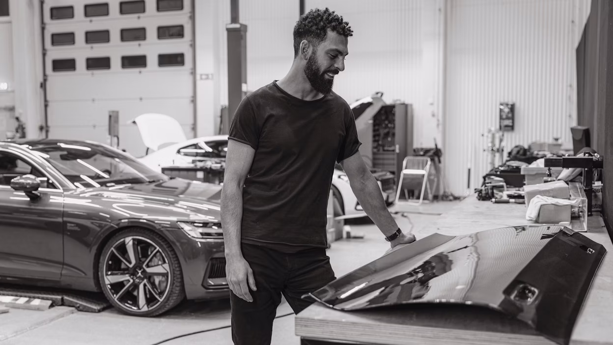 Person standing in a workshop near a Polestar car and a carbon fiber car panel on a table.