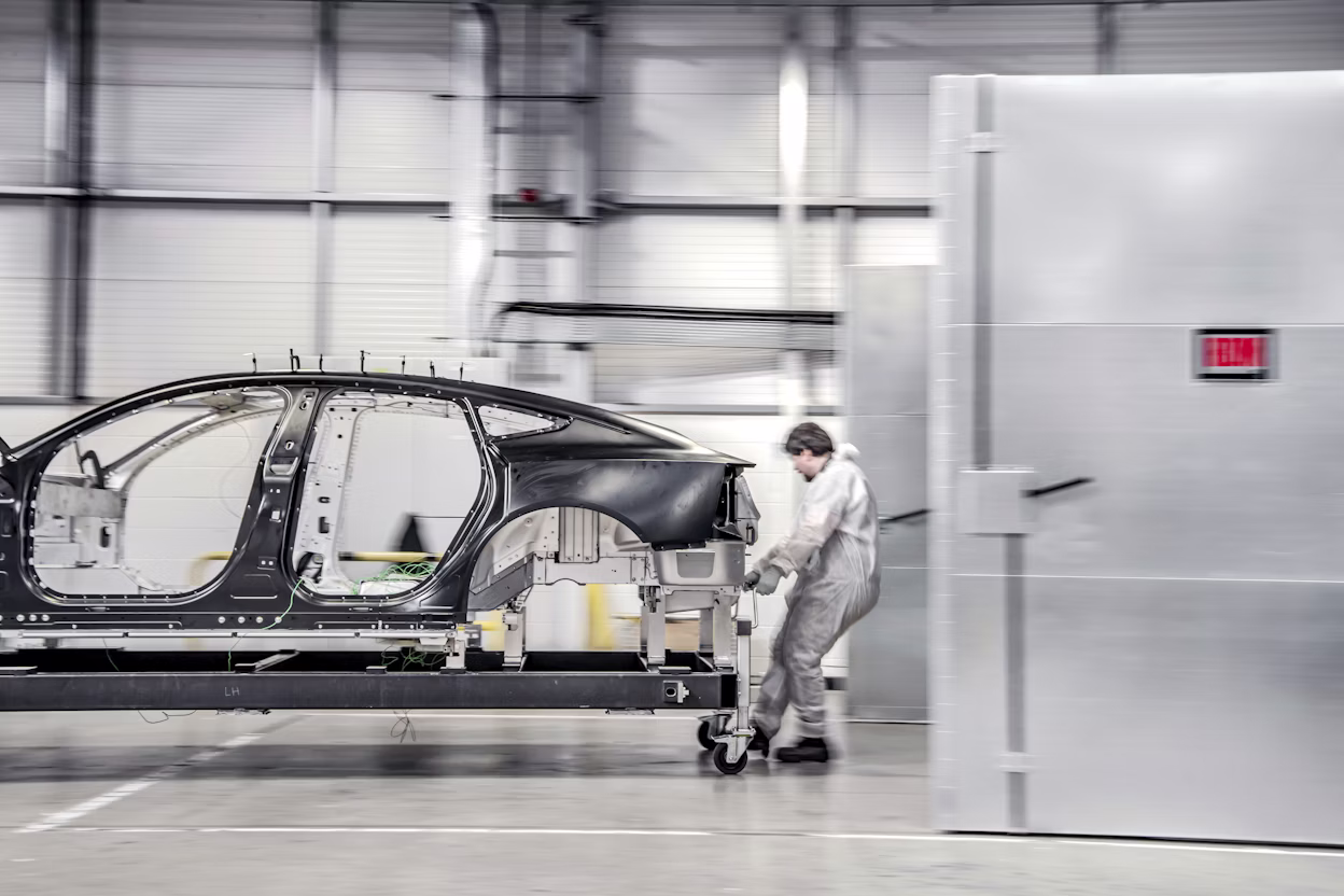 Worker pushes Polestar car frame on wheeled platform inside an industrial facility near a metal door labeled "Exit."