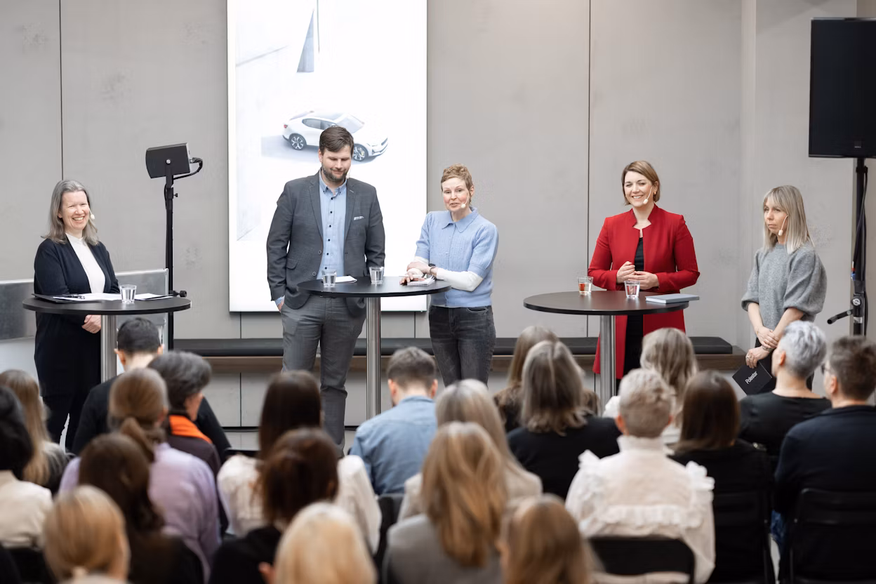 People on stage speak to an audience with a screen displaying a Polestar car in the background.