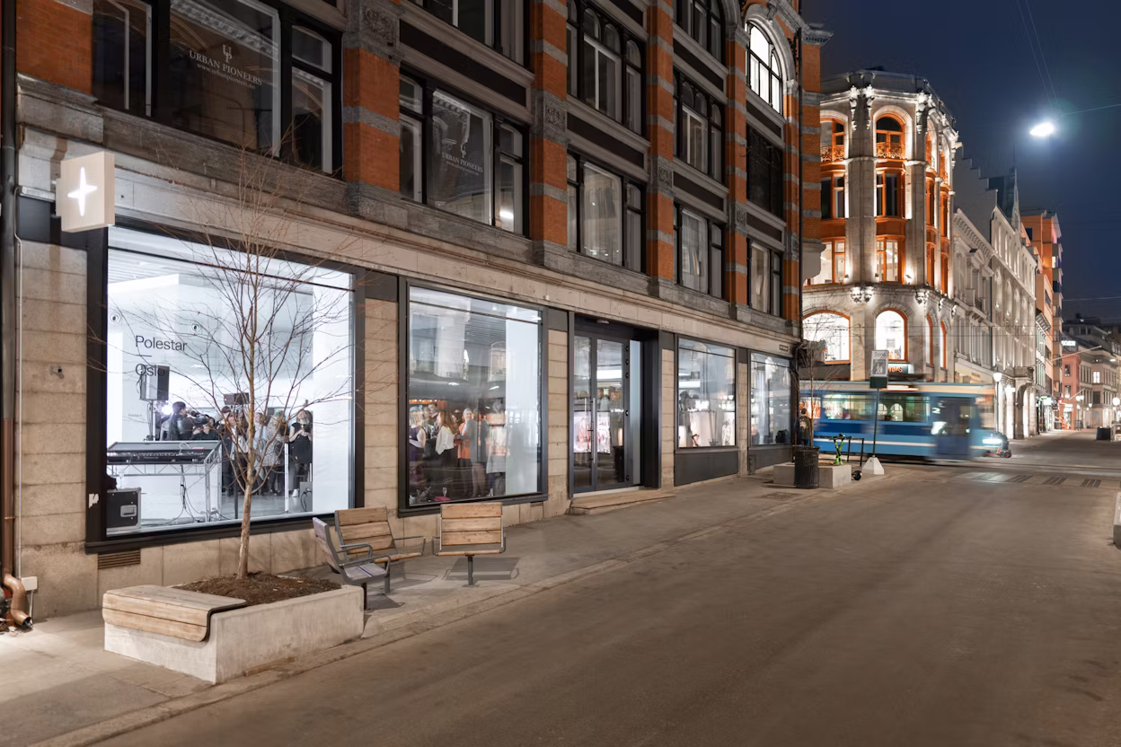 Street at night with a Polestar showroom in a building. Urban setting with benches, a tram, and illuminated building facades.
