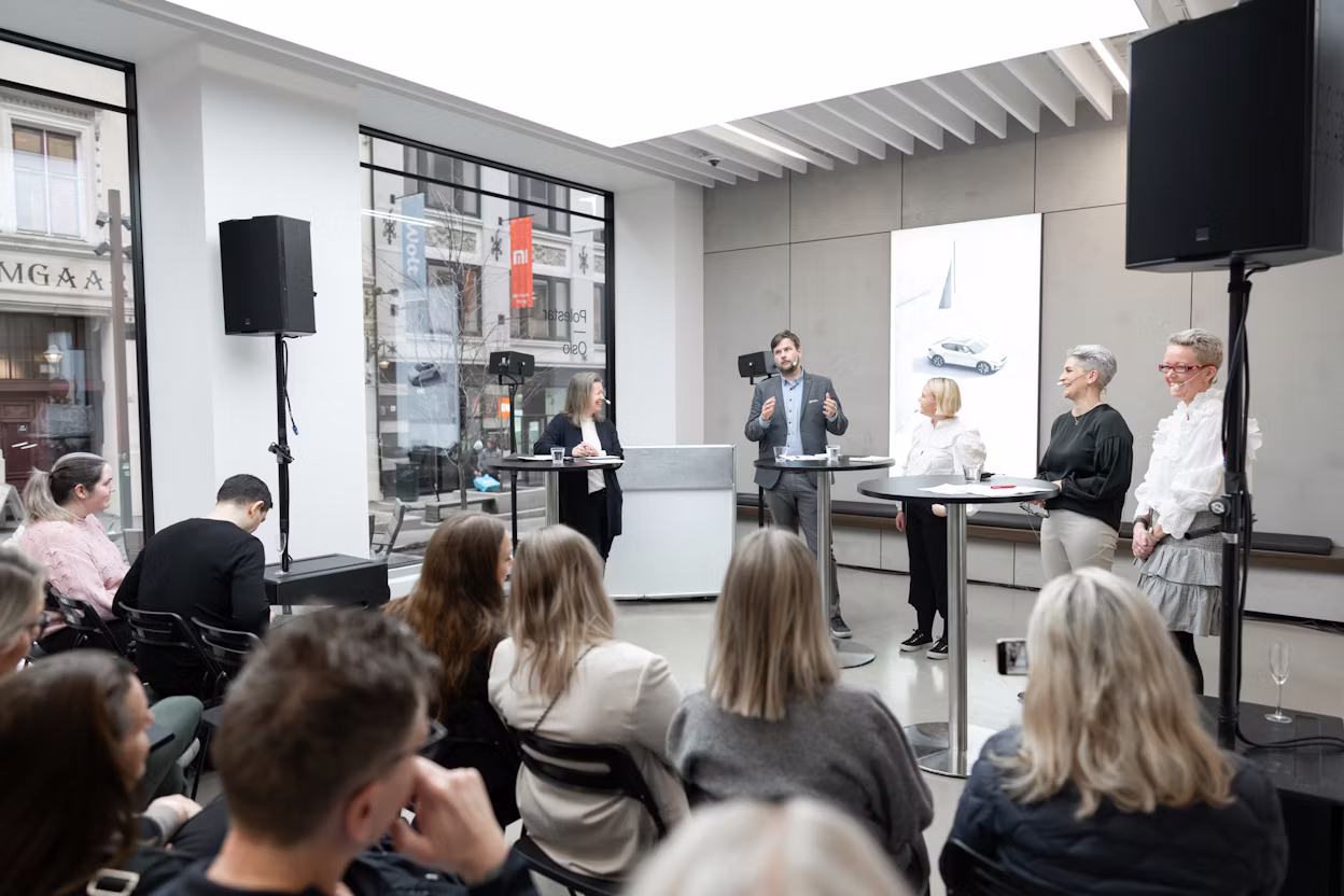 Panel discussion indoors with audience seated, speakers standing at tables, and a Polestar car poster displayed in the background.