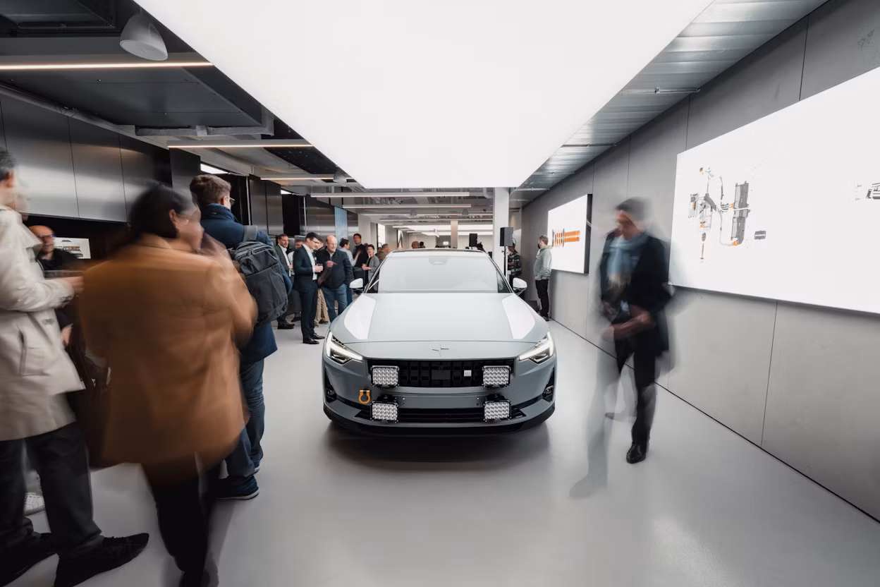 Polestar car displayed indoors surrounded by people and lit informational screens on the walls.