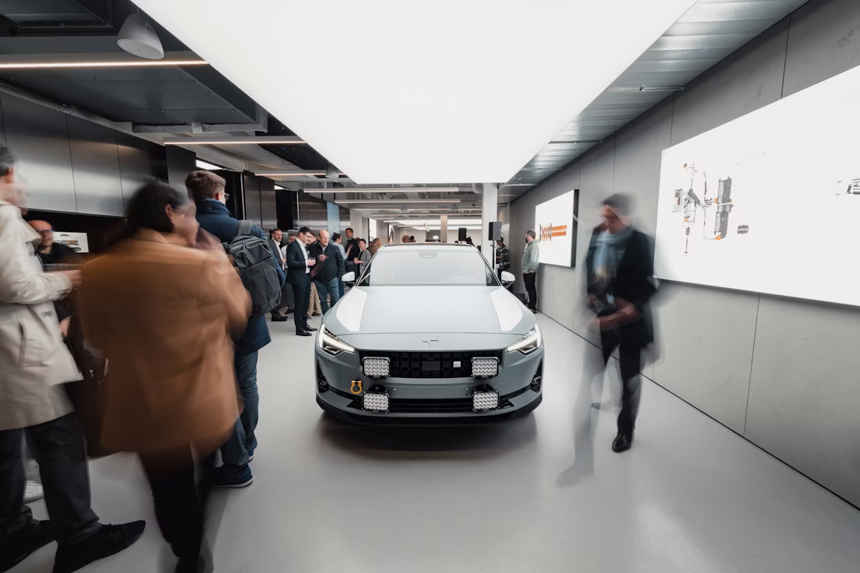 Polestar car displayed indoors surrounded by people and lit informational screens on the walls.