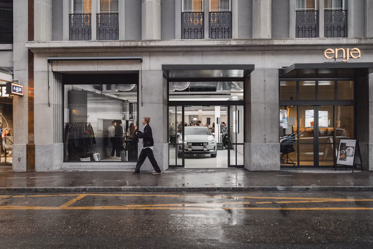 Storefront with Polestar car visible through glass doors, people inside, and a person walking past on a wet street.