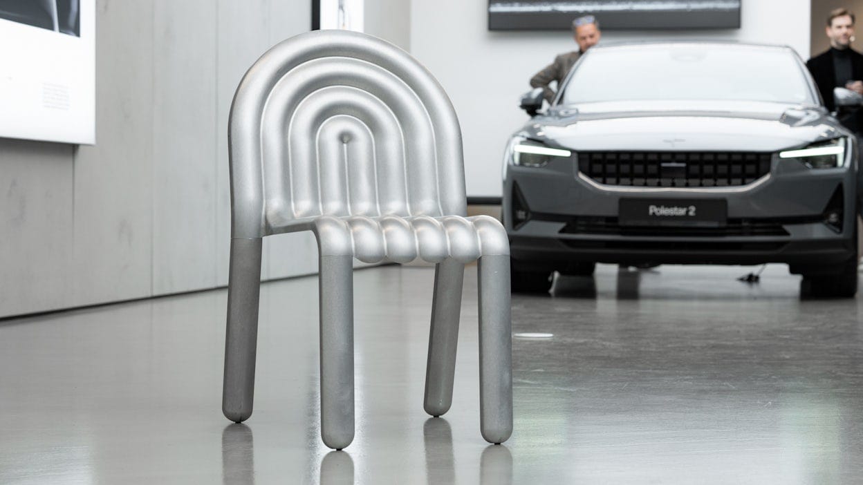 Image of an aluminium chair in front of a Polestar 2. 