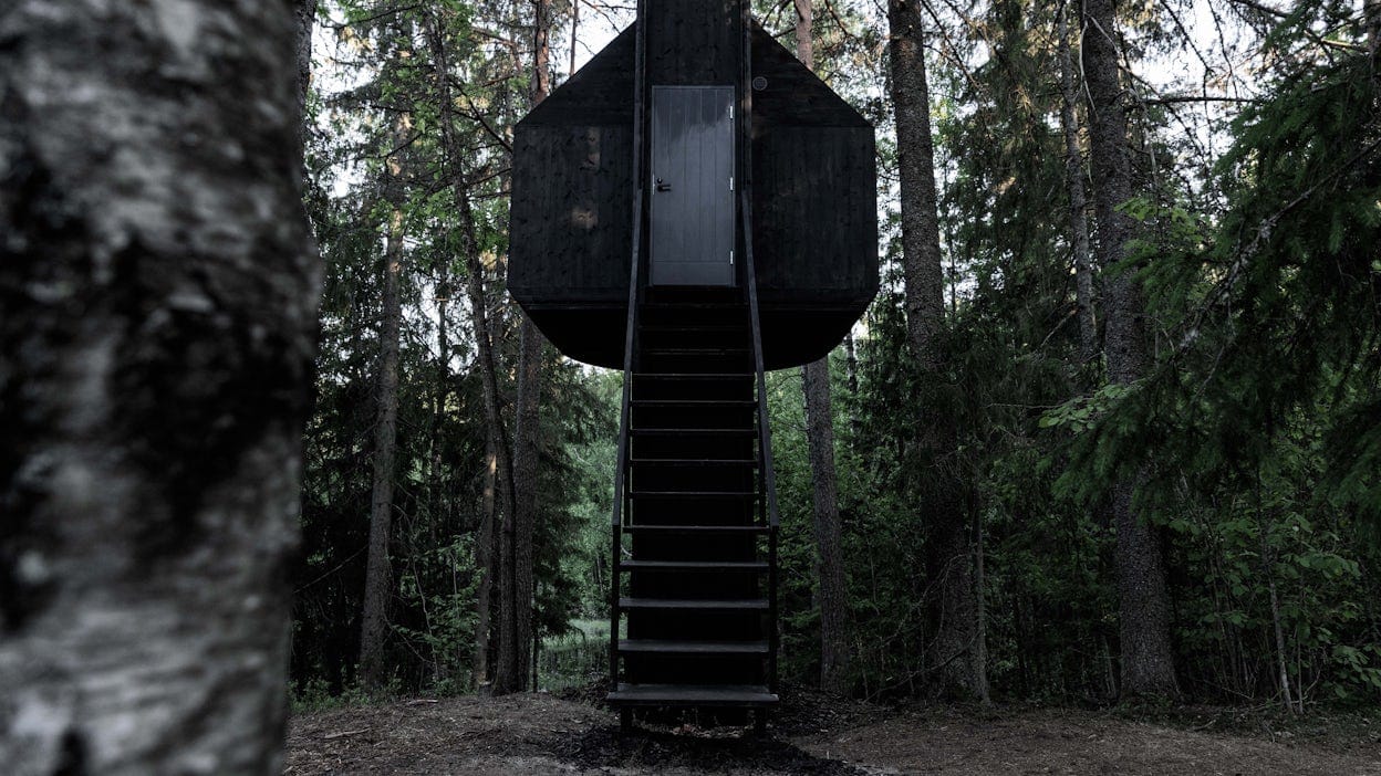 The exterior of a black treehouse with stairs leading up to an entrance. 