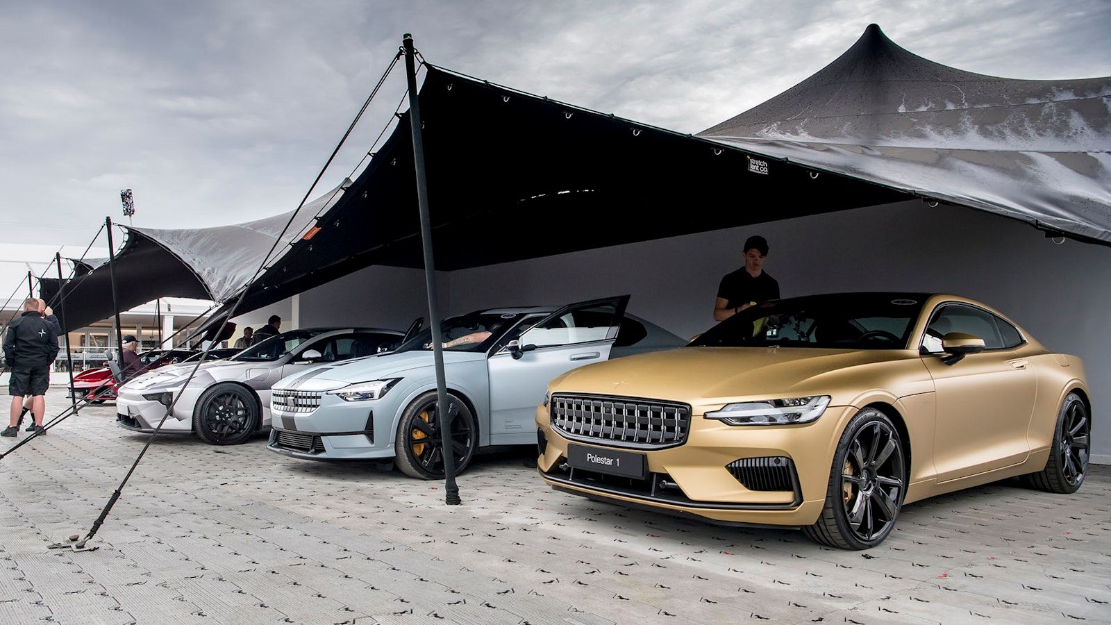 A camouflaged Polestar 5 prototype, light grey Experimental Polestar 2, and gold Polestar 1 parked under a cover.