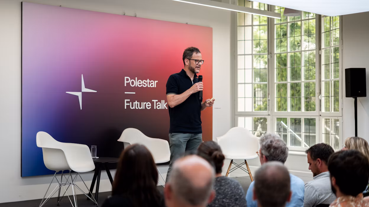 Speaker holding microphone on stage at Polestar Future Talk event with chairs, audience, and large windows in background.