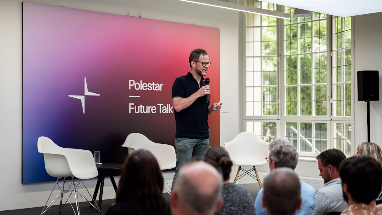 Speaker holding microphone on stage at Polestar Future Talk event with chairs, audience, and large windows in background.