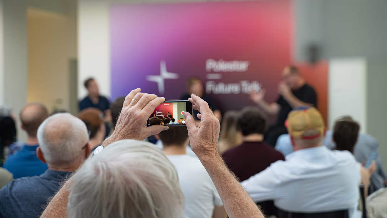Audience at Polestar Future Talk event, person holding phone capturing speakers on stage with Polestar logo in background.