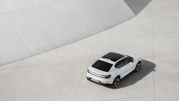 Aerial shot of the Polestar 2 electric vehicle showing the rear, right side profile and the top in which you can see the panoramic roof. The car is parked on large neutral tiles and casting a strong dark shadow in contrast to the car's bodywork, which is white.