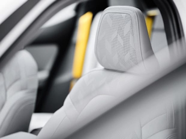 Interior shot of the Polestar 2 electric vehicle from through the front left side door window. The headrest and upper portion of the grey WeaveTech seat is shown, with contrasting yellow seat belts visible in the background.