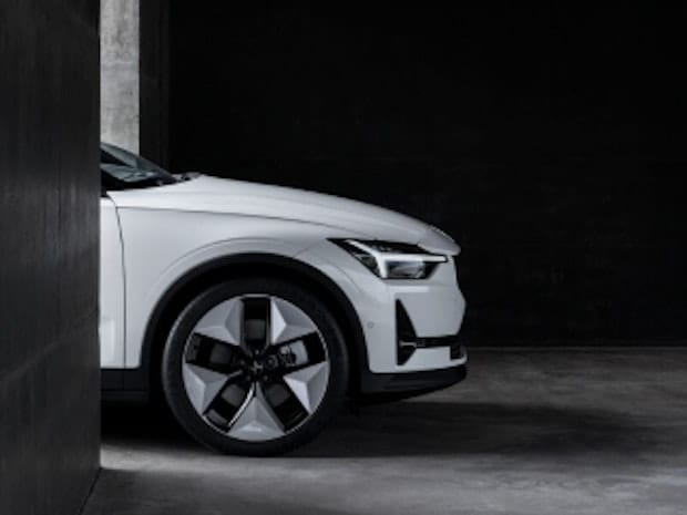 Right side front profile of the Polestar 2 electric vehicle, showing the front left wheel and headlights. The car is parked in a minimal grey concrete structure with the background in dark shadow, contrasting with the car's bodywork, which is white.
