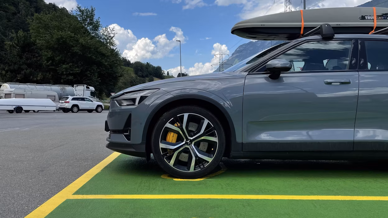 Polestar car parked on green pavement with roof box, surrounded by trees, hills, and other vehicles in the background.