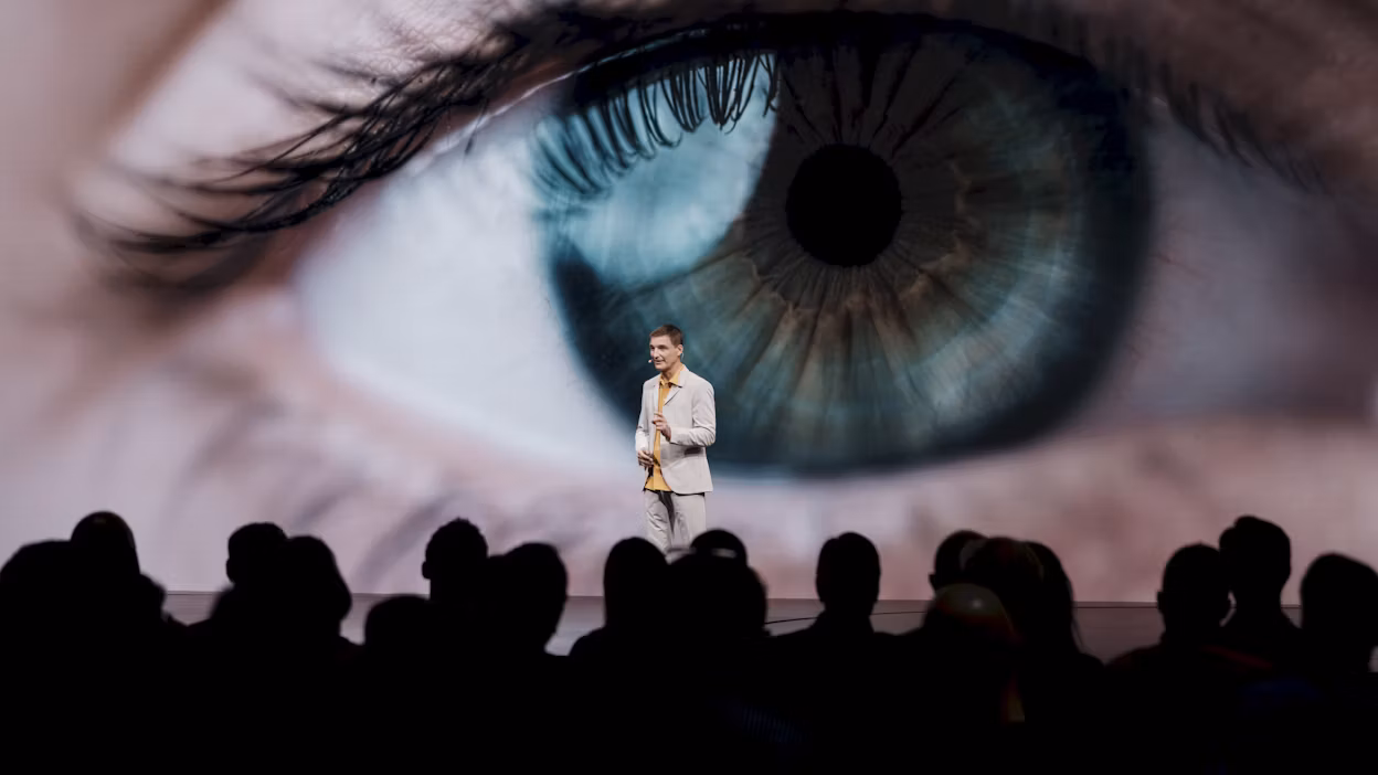 A man standing on stage in front of a screen showing a human eye.