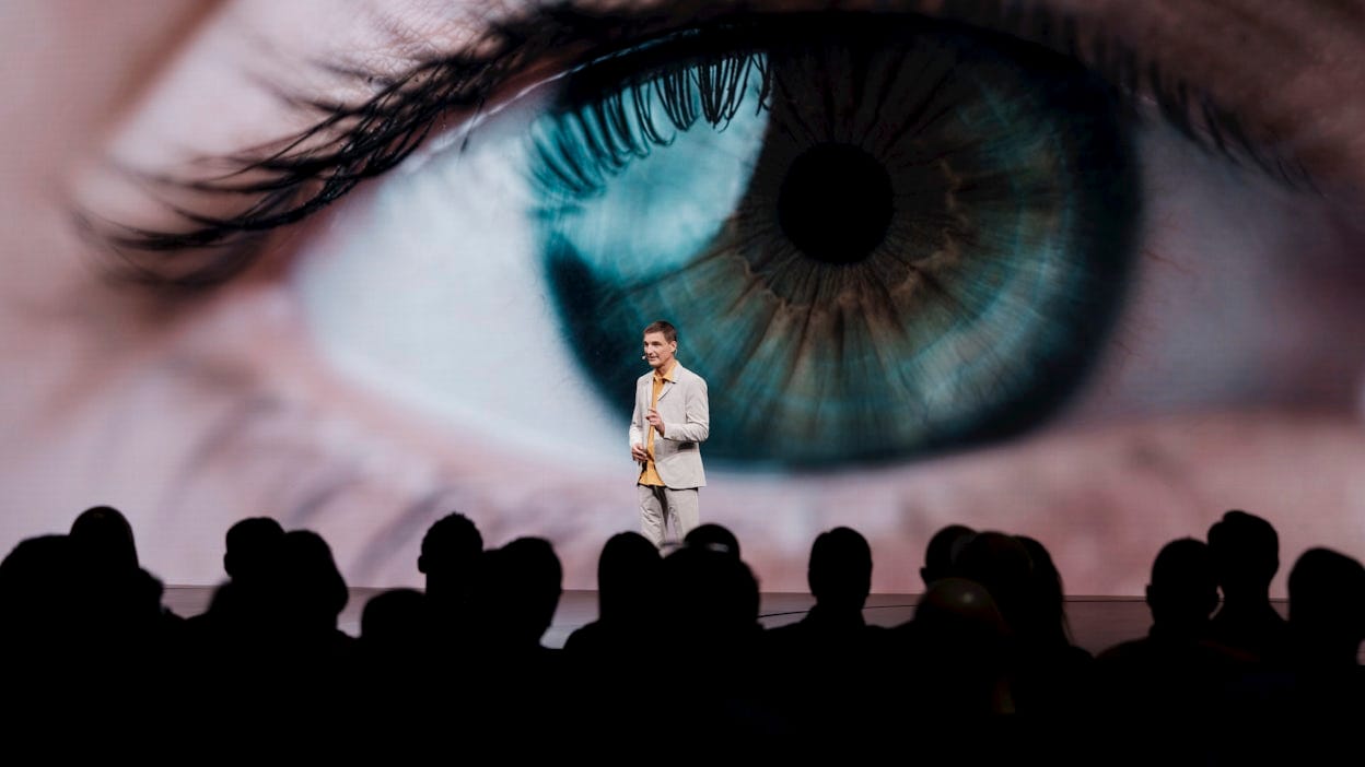 A man standing on stage in front of a screen showing a human eye.