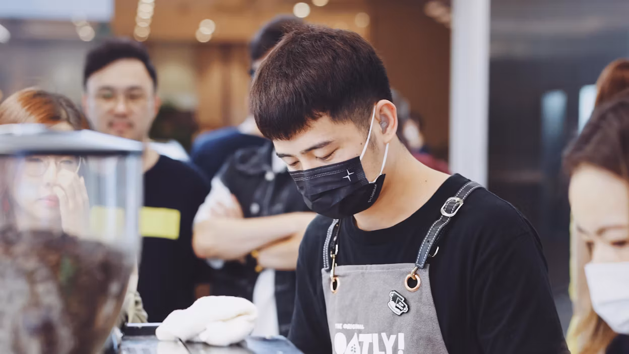 Barista wearing face mask with Polestar logo and apron with the Oatly logo.