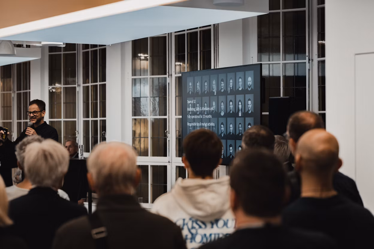 Audience watching a presentation with a screen displaying team information and photos in a room with large windows.