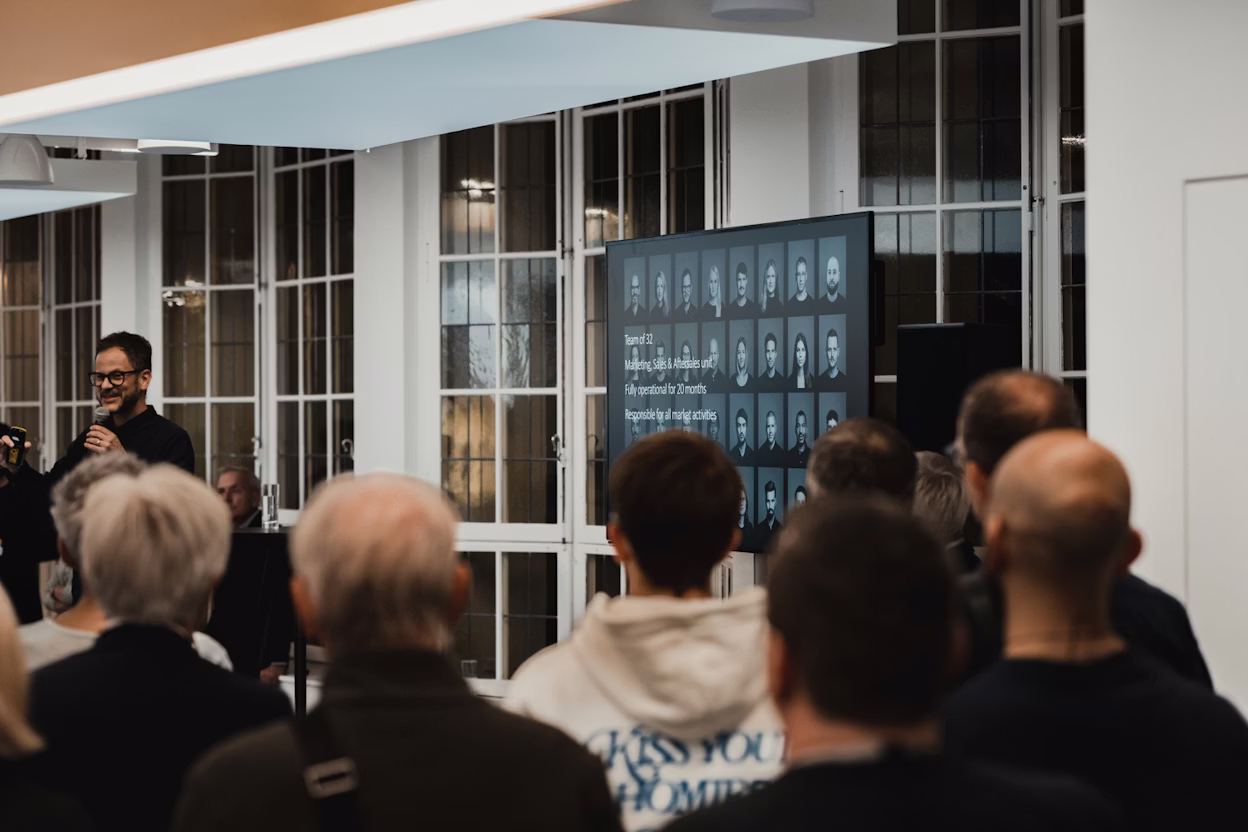 Audience watching a presentation with a screen displaying team information and photos in a room with large windows.