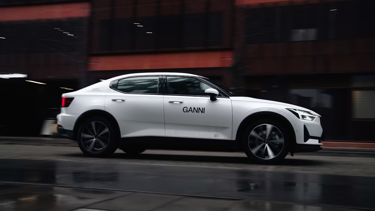 Side view of a white Polestar 2 with the Ganni logotype driving on a wet road.