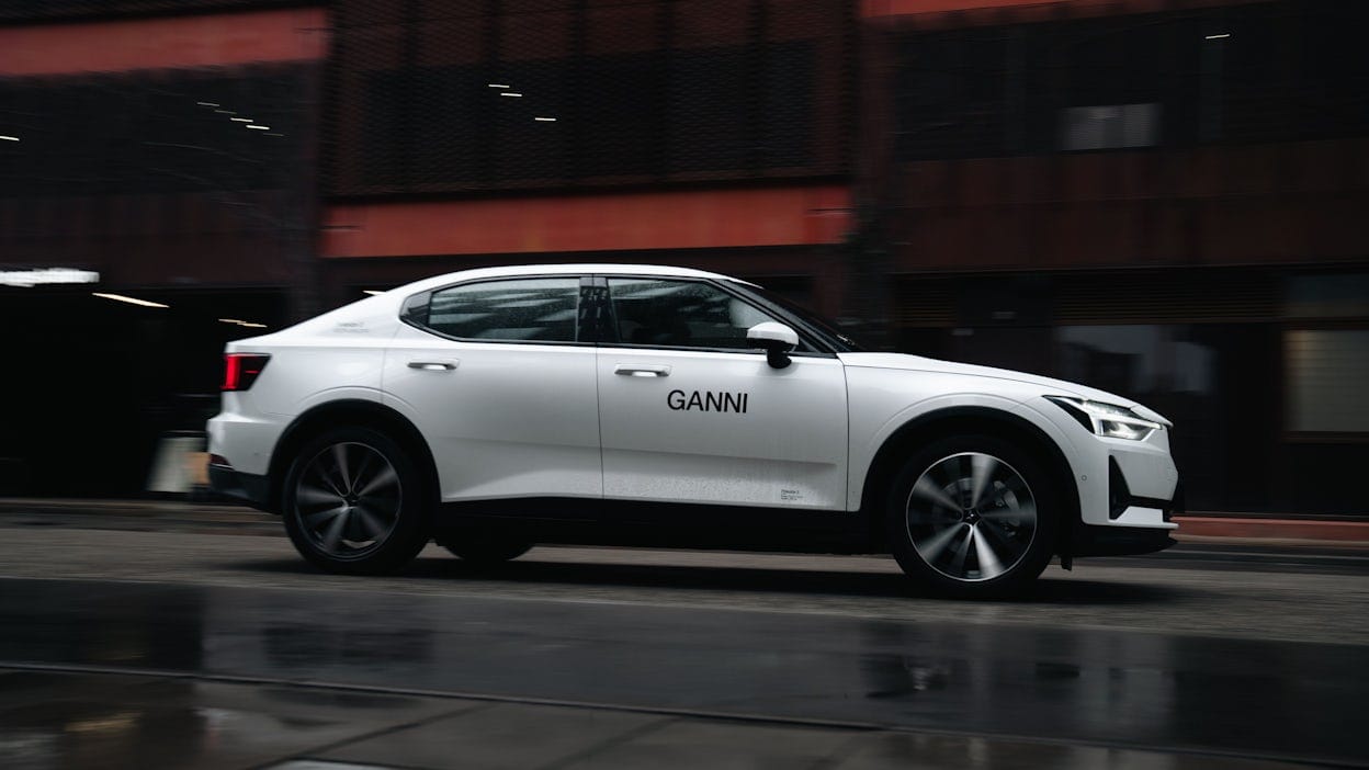 Side view of a white Polestar 2 with the Ganni logotype driving on a wet road.