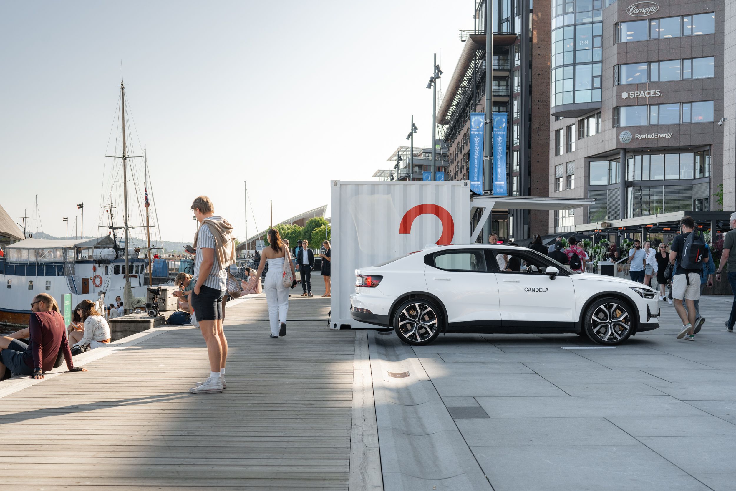 Polestar car parked near a waterfront with people walking and sitting, buildings and boats visible in the background.