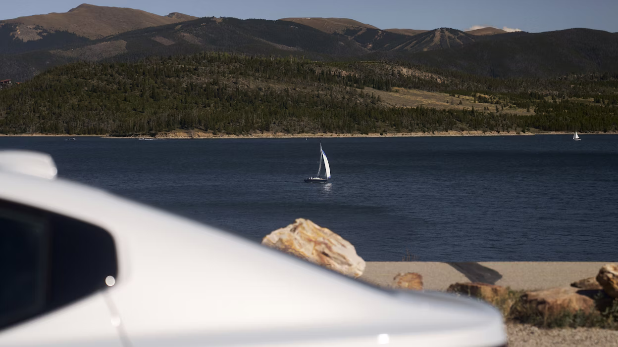 Polestar car in foreground, lake with sailboat and forested hills in background.