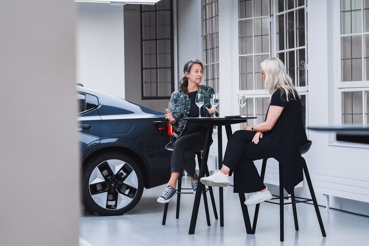 Two people seated at a table with glasses, Polestar car parked indoors near large windows in the background.